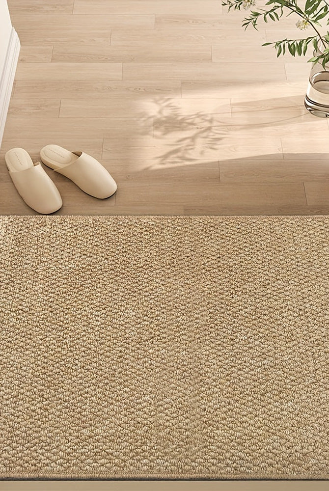 Beige doormat on a wooden floor with white slippers and a plant in the background