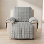 A light gray quilted slipcover with a textured grid pattern draped over a cream-colored recliner chair, set against a neutral beige wall and light wood floor.