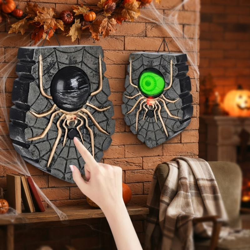 Two Halloween spider-themed wall decorations hanging on a brick wall, with one showing a black spider and the other showing a glowing green eye. A person's hand is touching the button on the left decoration, and a cozy fireplace scene with autumn decorations is blurred in the background.