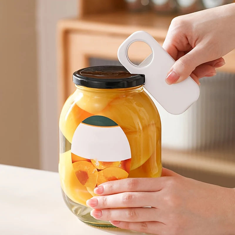 A person's hands are shown using a white plastic jar lifter tool to lift a heavy glass jar filled with sliced peaches, set against a softly blurred kitchen background.