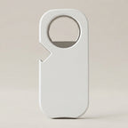 A minimalist white bottle opener with a clean, rounded rectangular design and a built-in circular cutout, standing upright on a neutral beige background.