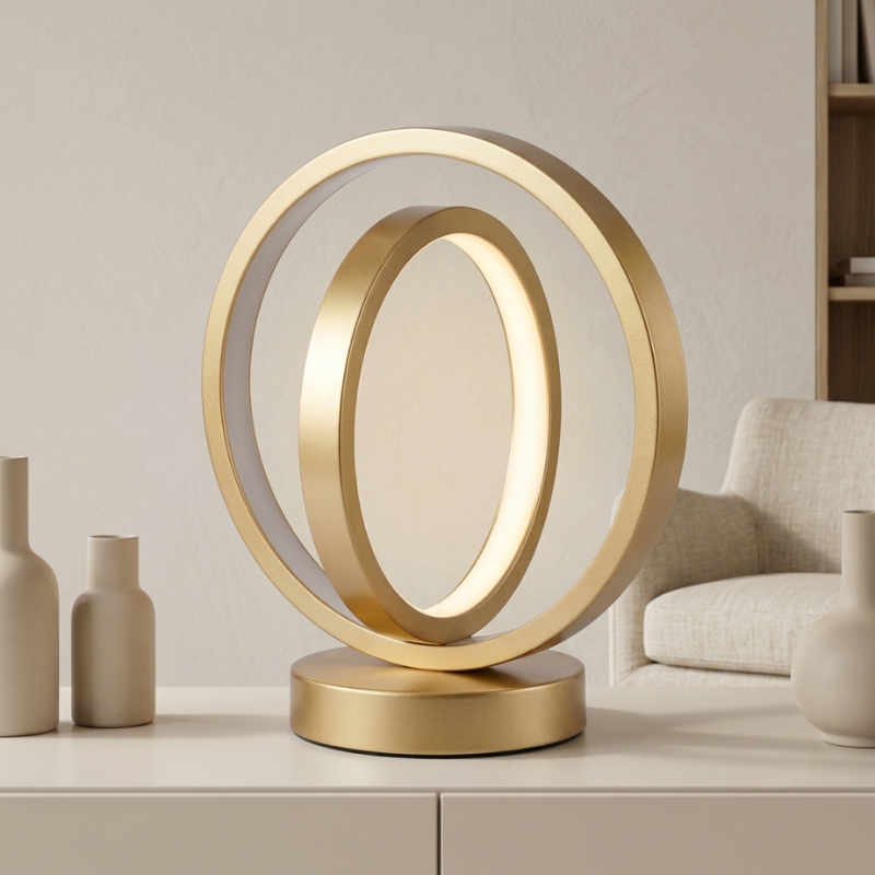 A modern gold-toned table lamp with two interlocking circular rings and a round base, shown lit on a white surface in a styled interior setting with neutral decor and a chair in the background.