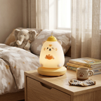 A cute, bear-shaped night light glowing with a warm yellow light on a wooden bedside table in a cozy bedroom. The light features a white bear face with orange details and a small yellow hat.