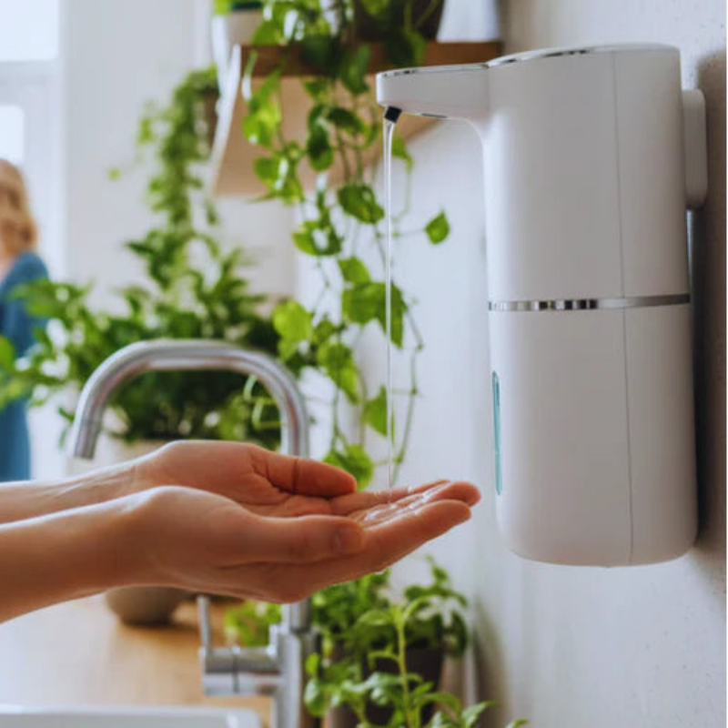 Hand using a white wall-mounted soap dispenser with plants and a kitchen sink in the background.