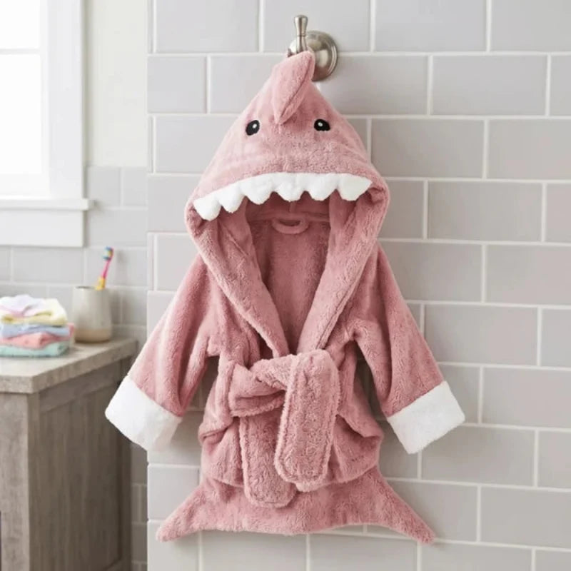 A pink plush shark-themed hooded bathrobe hanging on a wall hook in a bathroom setting. The robe features a shark head hood with embroidered eyes, white teeth, a tie belt, and white cuffs.