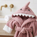 A pink plush hooded bathrobe designed like a shark, with white faux-teeth along the mouth, black eyes, and white cuffs, styled on a bathroom counter next to a rolled white towel.
