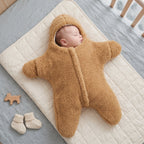 A newborn baby wearing a plush, star-shaped brown onesie sleep sack, lying on a cream quilted mattress. A pair of cream knit baby booties and a small wooden toy are placed nearby on a gray blanket.