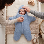 A baby wearing a light blue fuzzy hooded footed swaddle lies on a woven blanket in a crib, with a mother's hand touching the hood and a father's hand adjusting the swaddle. A small stuffed deer toy is visible at the bottom right.