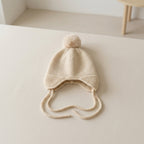 A cream-colored knit baby hat with a matching pom-pom and ear ties, displayed on a light beige surface.
