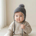 A baby wearing a gray knit pom-pom beanie hat with ear flaps and drawstrings, paired with a beige knit sweater, in a softly lit indoor setting.