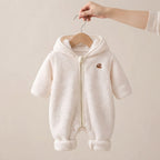 A baby hooded romper in soft cream fleece fabric, hanging on a wooden hanger held by a person's hand against a plain beige background.