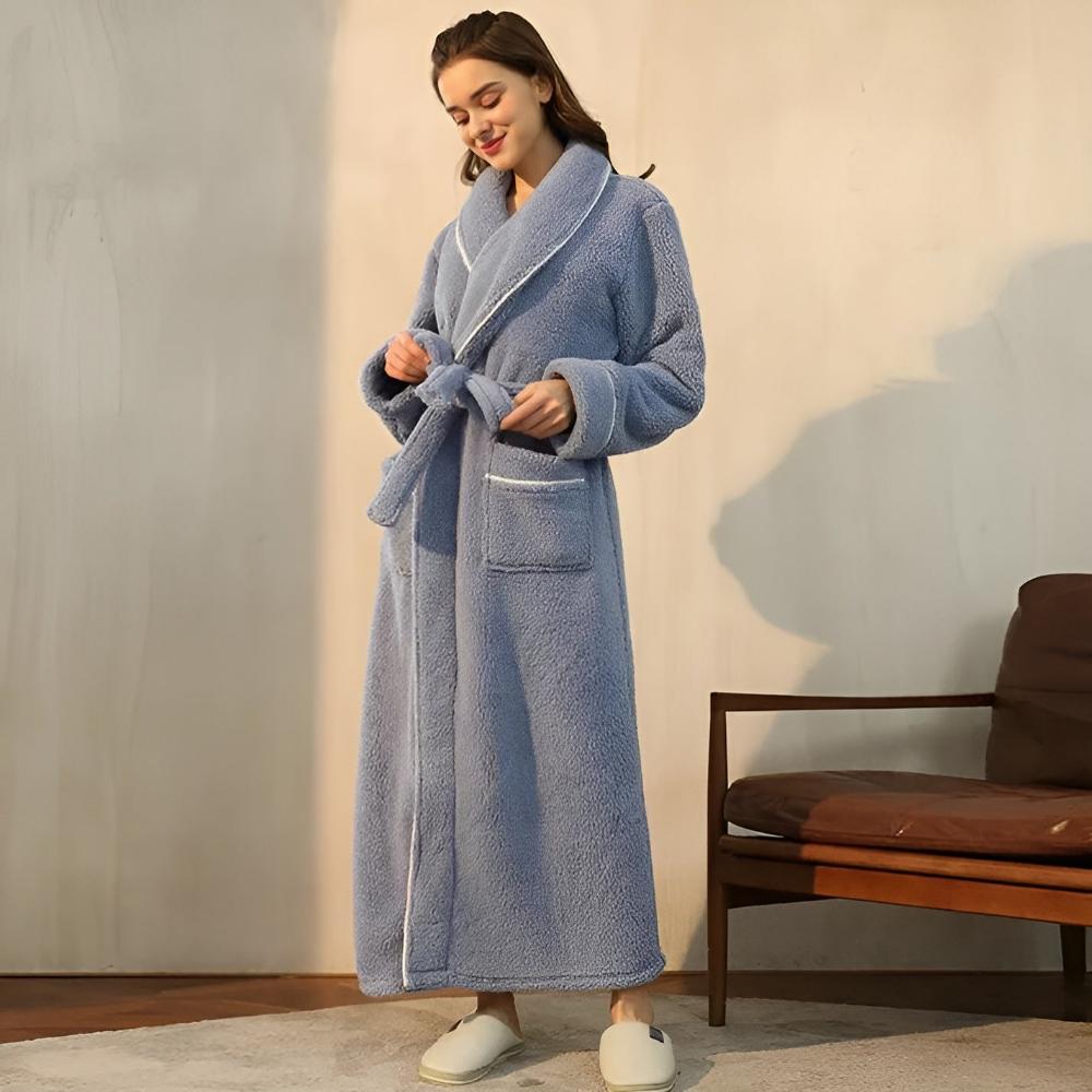 Woman wearing a blue robe standing in a room with a wooden chair and beige wall.