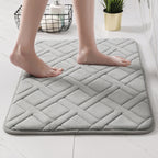 A light gray quilted bath mat with a diamond pattern, shown with bare feet standing on it in a bright bathroom setting.