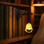 A small ghost-shaped wall lamp glowing warm yellow, holding a book, mounted on a black wrought-iron bracket attached to a wooden bookshelf in a cozy library setting.