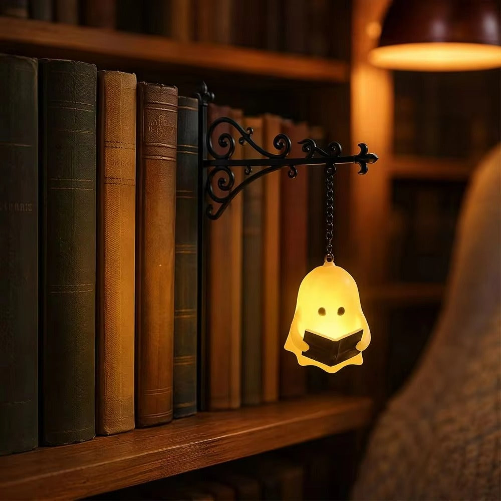 A small ghost-shaped wall lamp glowing warm yellow, holding a book, mounted on a black wrought-iron bracket attached to a wooden bookshelf in a cozy library setting.