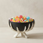 A black bowl filled with colorful lollipops and candy, supported by white skeletal hands on a stand, set against a neutral beige background.