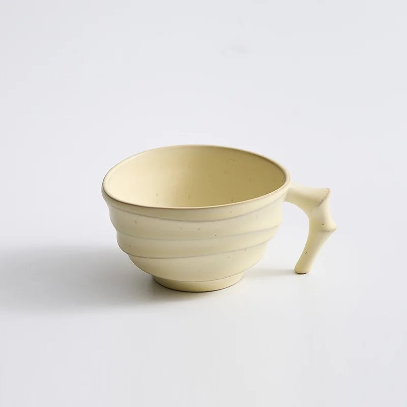 A pale cream-colored ceramic cup with a unique handle shaped like a small branch, set against a plain white background.