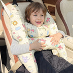 A smiling young boy sits in a car seat using a padded baby support pillow with a cream background and colorful cartoon animal prints.
