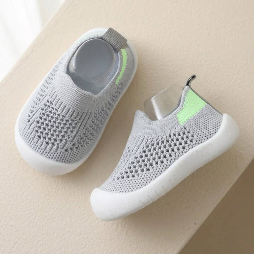 A pair of light gray knit slip-on baby shoes with white soles and a small neon green accent on the heel, displayed on a beige surface.