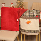 Two Christmas-themed fabric chair covers on beige dining chairs. One cover is solid red with a Santa hat design, and the other is white with a snowman face, plaid hat, and decorative trim.