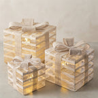 Three stacked gift box lanterns in different sizes, decorated with beige burlap ribbons and bows, glowing with warm internal fairy lights against a neutral beige background.