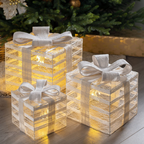 Three square decorative gift box lanterns with white textured wrapping and white ribbons, glowing with warm yellow lights, arranged on a wooden surface in front of a Christmas tree.