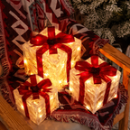 Three glowing Christmas gift box string lights with red ribbon bows, arranged on a patterned fringed blanket on a wooden chair.