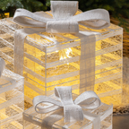 A decorative gift box lantern with a glowing warm yellow light inside, wrapped in a sheer white burlap ribbon and bow, shown on a softly blurred outdoor background.