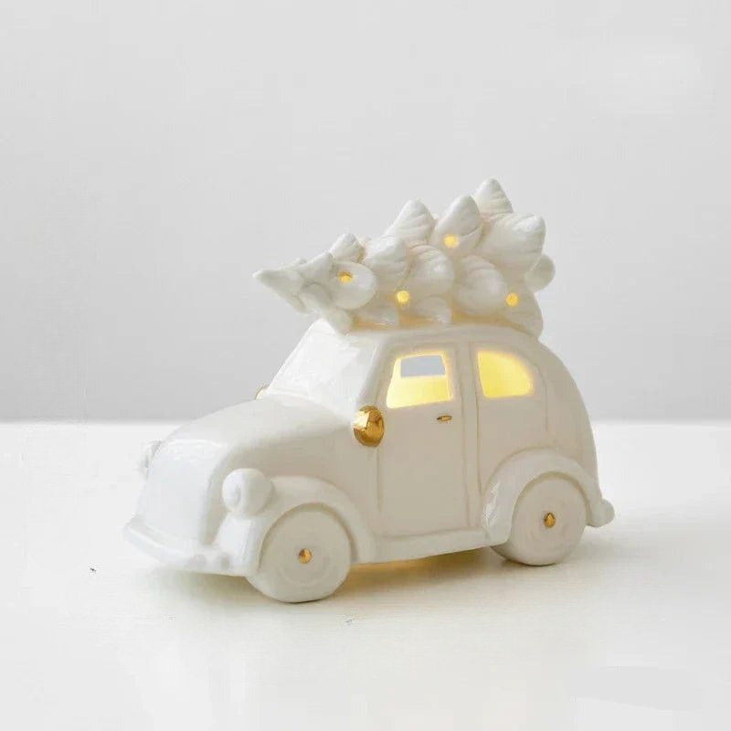 A white ceramic Christmas ornament shaped like a vintage car with a tree on its roof, featuring warm yellow lights glowing from within and small gold accents on the wheels and door.