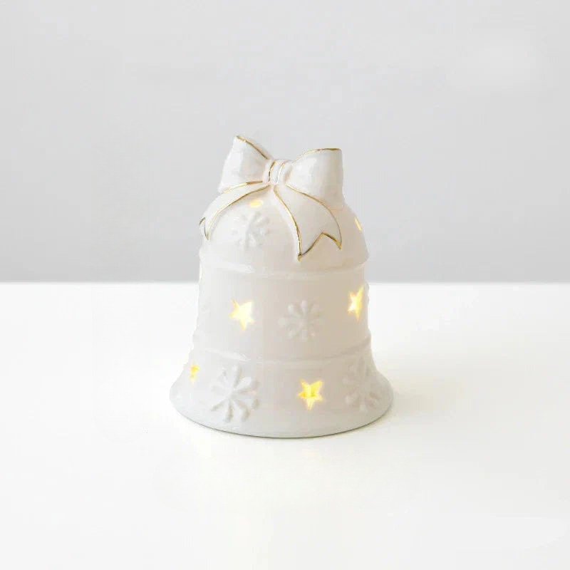 A white ceramic Christmas bell candle holder with a bow on top, featuring cutout snowflakes and stars that glow warm yellow light, set against a clean white background.