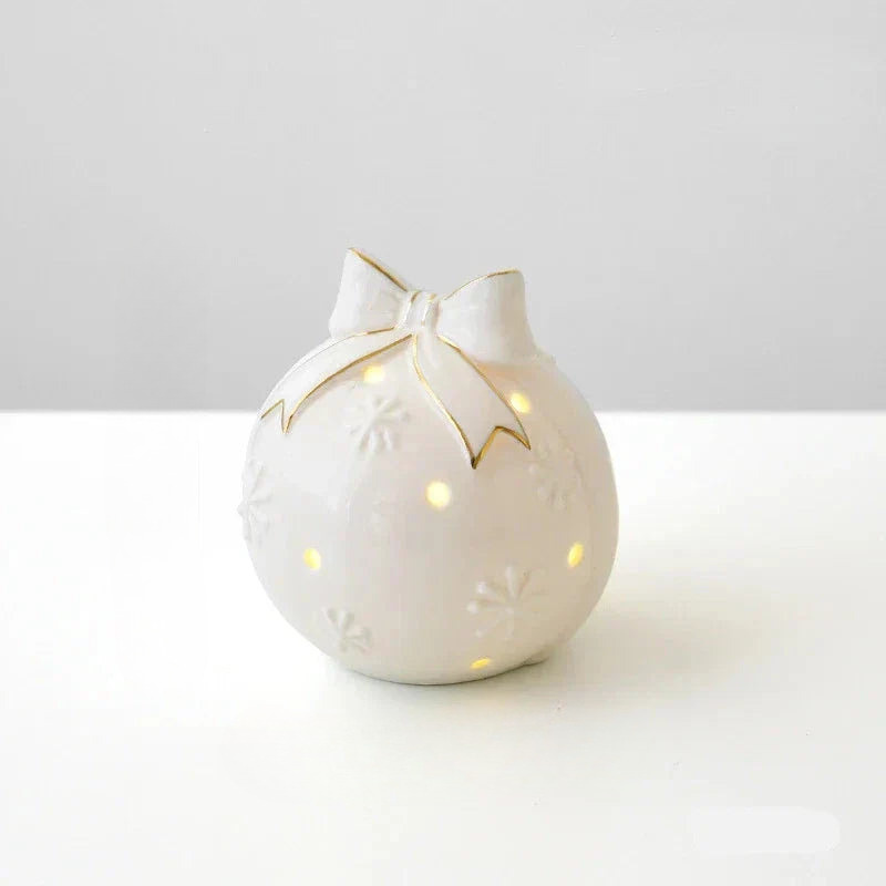 A white ceramic Christmas ornament-shaped lamp with a bow on top, featuring cutout details that reveal warm yellow light, set against a clean white and light gray background.