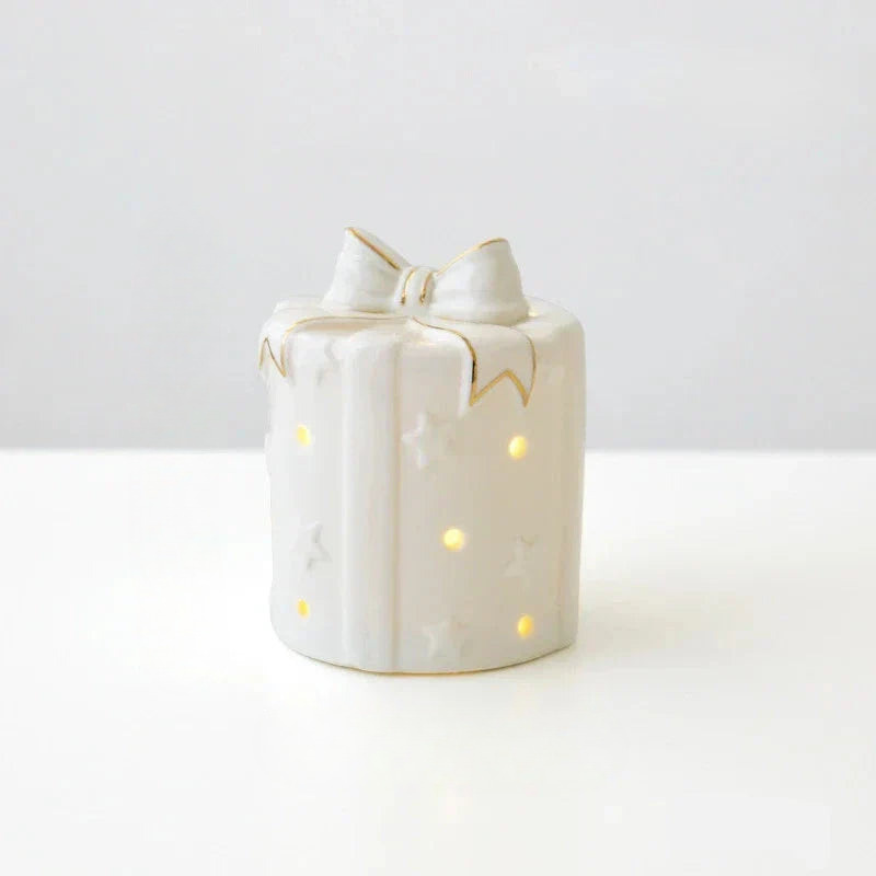 A small white gift box candle holder with a bow on top, featuring star and wave cutouts that glow with warm yellow light, set against a clean white background.
