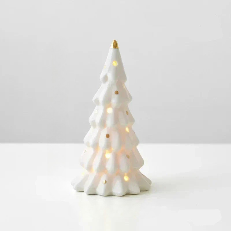 A small white ceramic Christmas tree figurine with gold accents and warm internal string lights, standing on a white surface against a light gray background.