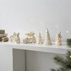 A set of white ceramic Christmas ornaments and figurines, including a snowman car with a Christmas tree on top, a small house, a snowman, a cone tree, and a snowman with warm glowing lights, arranged on a white shelf.