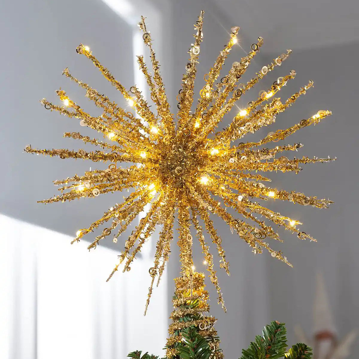 A gold metallic starburst Christmas tree topper with warm white LED lights, shown mounted on a gold tree trunk with green pine branches visible at the base against a soft gray indoor background.