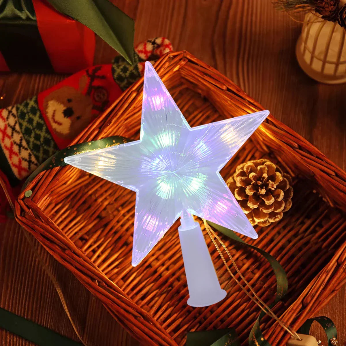 A glowing five-point star tree light with a white plastic base, displayed on a wicker basket with holiday decorations including a pinecone and ribbons.