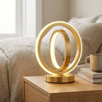 Gold circular table lamp on a wooden nightstand with a bed in the background