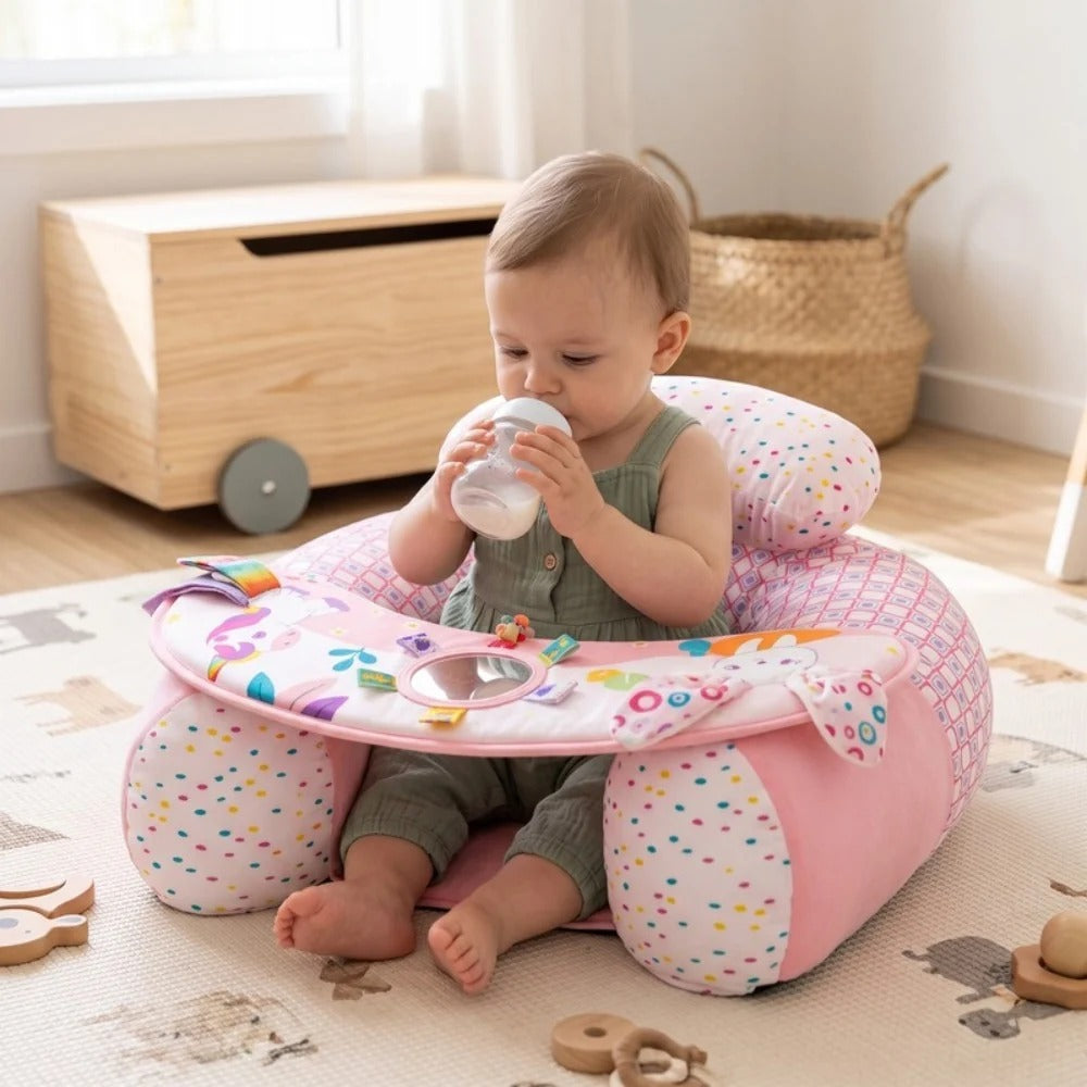 A baby sitting in a pink patterned baby seat on a play mat, drinking from a bottle. The seat has colorful animal and star patterns, and the room has a wooden toy chest and basket in the background.