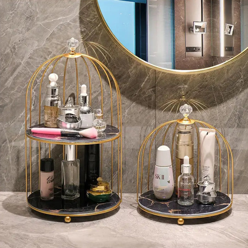 Two gold birdcage-style cosmetic organizers with marble trays are shown on a bathroom counter in front of a round mirror. The organizer holds assorted skincare and beauty products.
