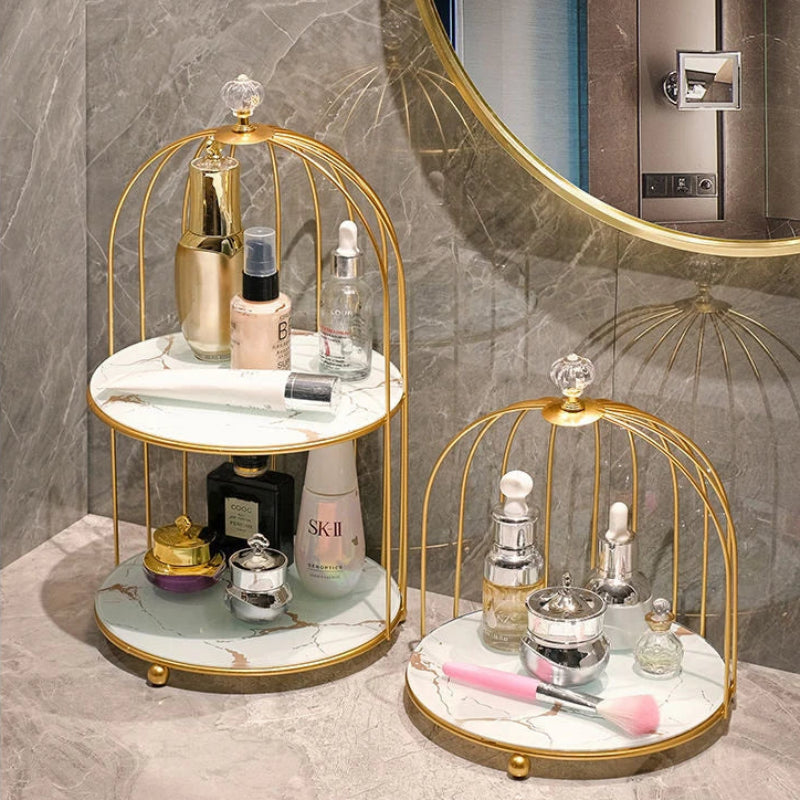 Two round two-tier bathroom vanity stands with gold metal birdcage frames and white marble-look shelves are shown holding assorted skincare and perfume bottles. The larger stand stands on a marble countertop against a gray marble wall, with a round mirror partially visible behind it.