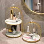 Two round two-tier bathroom vanity stands with gold metal birdcage frames and white marble-look shelves are shown holding assorted skincare and perfume bottles. The larger stand stands on a marble countertop against a gray marble wall, with a round mirror partially visible behind it.