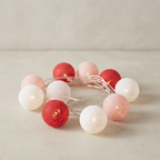A string of decorative globe fairy lights in white, light pink, and coral red, arranged in a circle on a neutral beige surface with a matching background.
