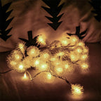 Warm white LED fairy string lights with fluffy pom-pom bulbs, shown illuminated on a dark surface against a dimly lit background with black tree silhouettes.