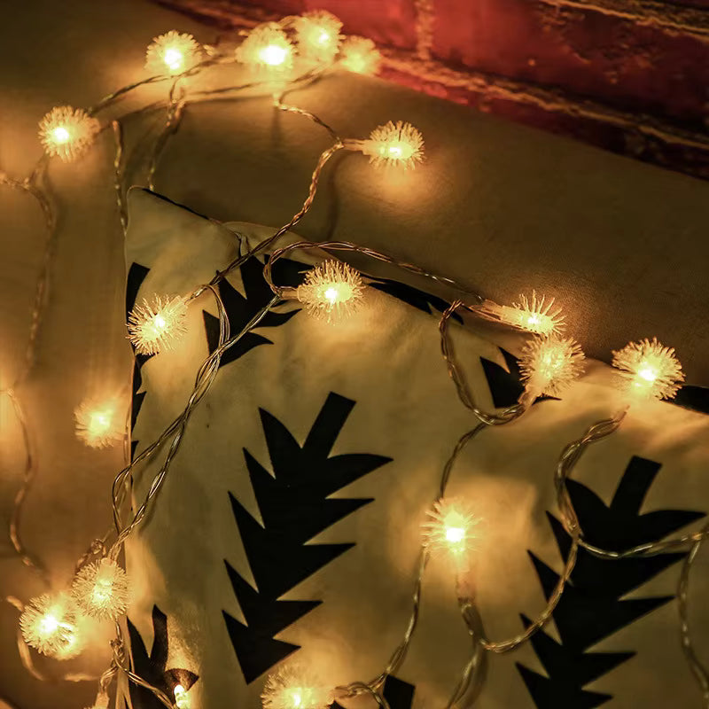 Warm white LED string lights with fluffy ball-shaped bulbs draped over a fabric backdrop with black leaf cutouts, against a rustic red-brown background.