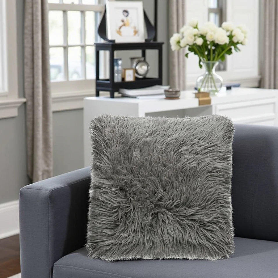 Gray fluffy pillow on a gray sofa in a living room setting with a vase of flowers and a shelf in the background.