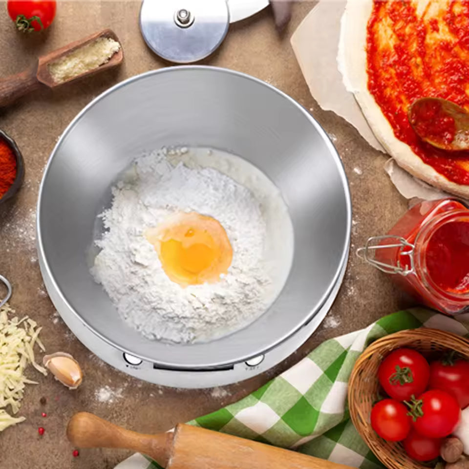 A stainless steel planetary mixer bowl sits on a countertop, filled with flour and a raw egg yolk, surrounded by pizza ingredients including a pizza crust, tomatoes, garlic, cheese, and a rolling pin.