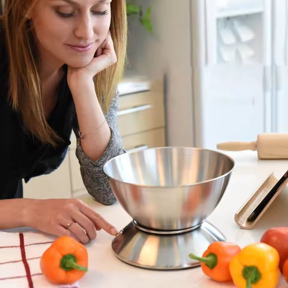 A woman in a kitchen looks down at a stainless steel countertop scale with a mixing bowl on top, surrounded by colorful bell peppers.