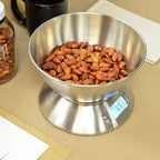 A stainless steel digital food scale with a bowl filled with almonds, shown on a woven beige mat with a black mug and a jar in the background.