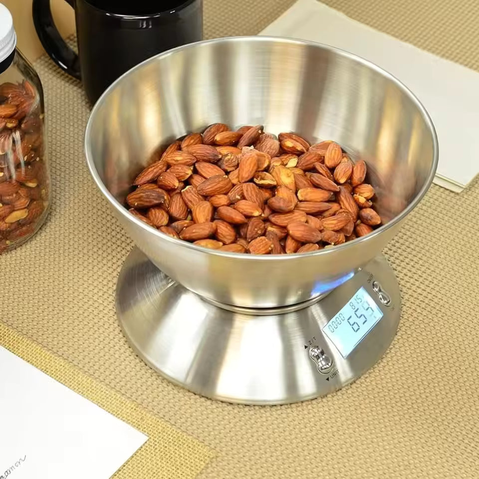 A stainless steel digital food scale with a bowl filled with almonds, shown on a woven beige mat with a black mug and a jar in the background.