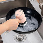 Hand using a handheld scrubber to clean a black pan in a sink.